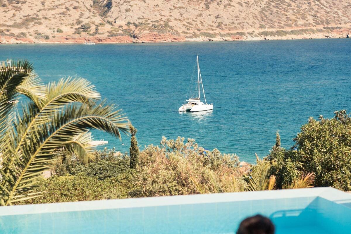 A person sitting by a pool with a view of a sailboat on the water and a mountainous landscape in the background.