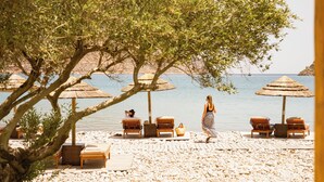Een strand met houten ligbedden, rieten parasols en een rotsachtige kustlijn.