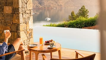 Una persona descansando en una silla de mimbre junto a una mesa de madera con una bebida, con vistas a un lago con un bote y montañas al fondo.