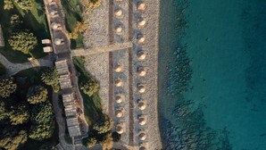 Luchtbeeld van een kustresort met houten huisjes langs het strand, een verhard pad en een rotsachtige kustlijn.