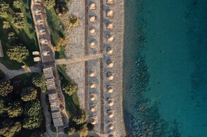 An aerial view of a coastal resort with wooden cabins aligned along a beachfront, a paved walkway, and a rocky shoreline.