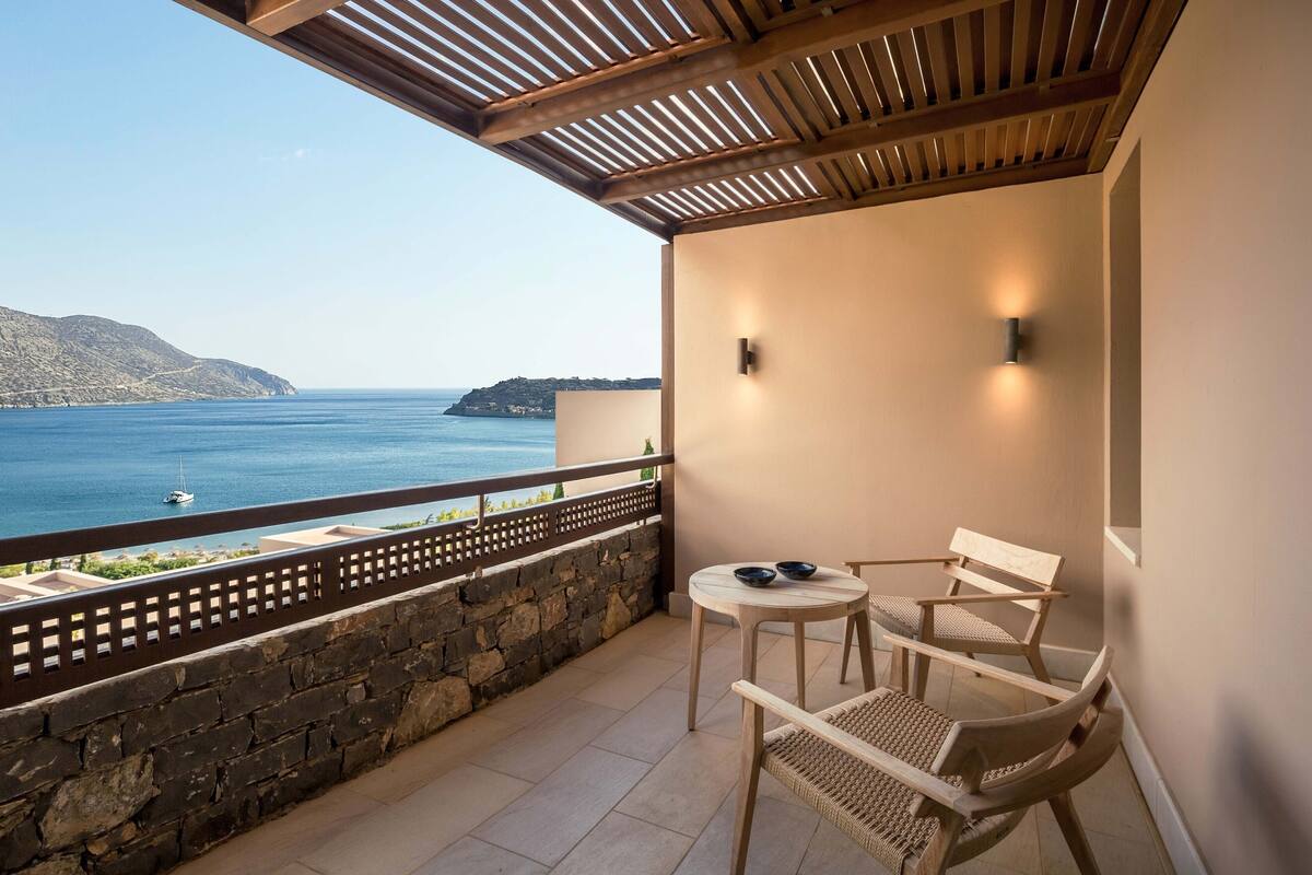 A balcony with a view of the sea, stone wall, and two wooden chairs.
