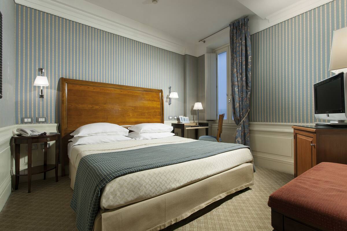 Ein Hotelzimmer mit einem großen Bett, Nachttischen, einem Schreibtisch, einem Fernseher und einem Fenster mit Vorhängen.