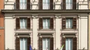 Un edificio de varios pisos con fachada simétrica, ventanas uniformemente distribuidas con contraventanas, una entrada central con toldo y banderas en el segundo piso.
