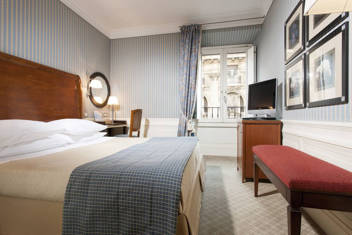 Ein Hotelzimmer mit einem großen Bett, einem Nachttisch mit Lampe, einem Fernseher, einer Bank und einem Fenster mit Blick auf ein Gebäude.