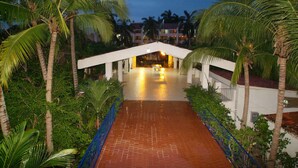A covered walkway with a tiled floor, flanked by palm trees and greenery, leading to a building with a white facade and a sloped roof.