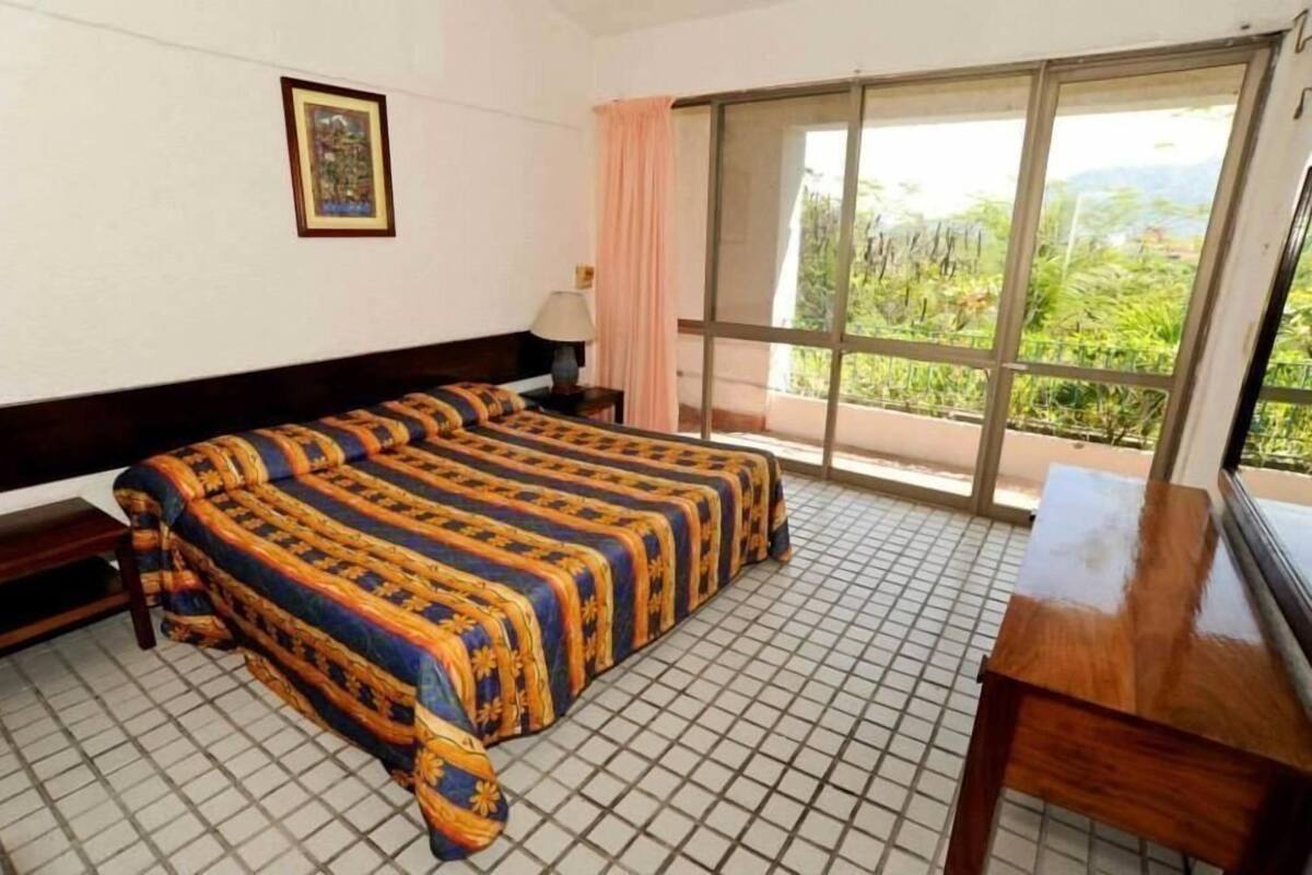 Habitación de hotel con una cama grande, un escritorio, un ventanal con vistas a zonas verdes y un cuadro en la pared.