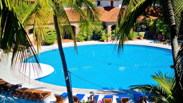A large, clear blue swimming pool surrounded by lounge chairs and palm trees, with a building in the background.