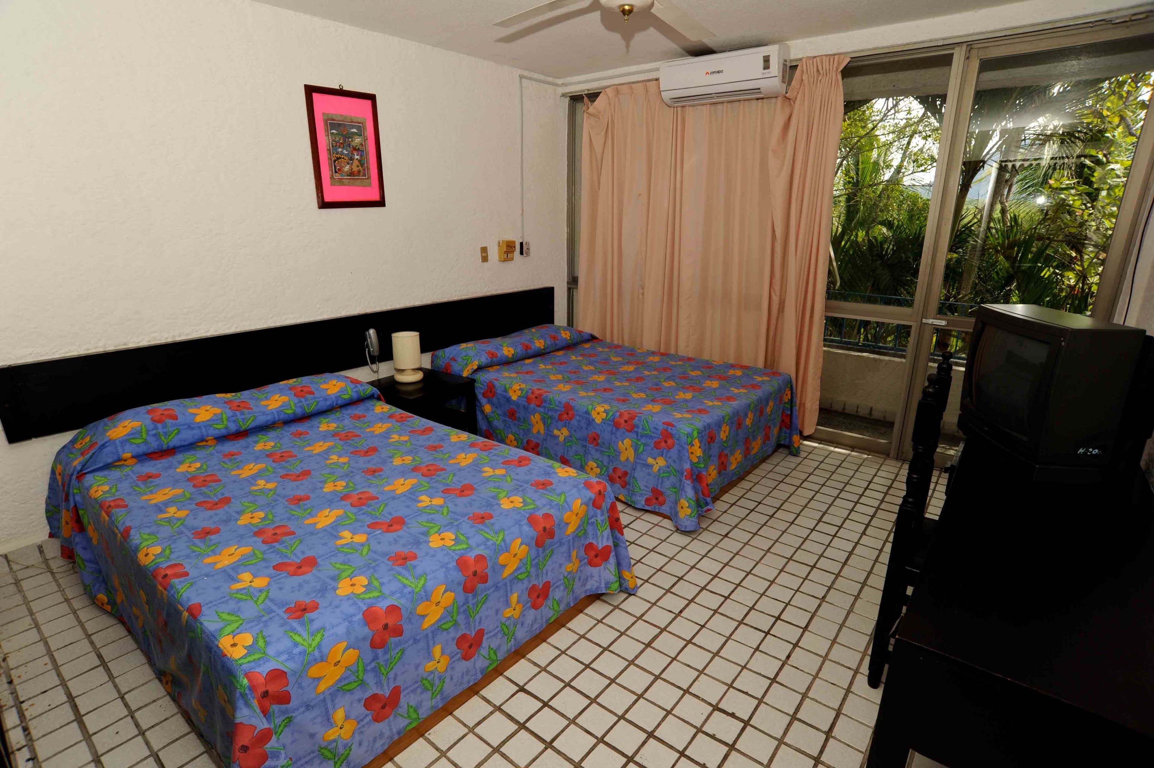 Habitación de hotel con dos camas, ventilador de techo, aire acondicionado, televisión y ventana con cortinas.
