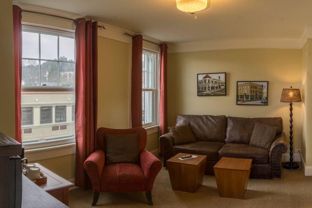 A living room with a red armchair, a brown leather sofa, a wooden coffee table, and a floor lamp. There are two framed pictures on the wall and a window with red curtains.