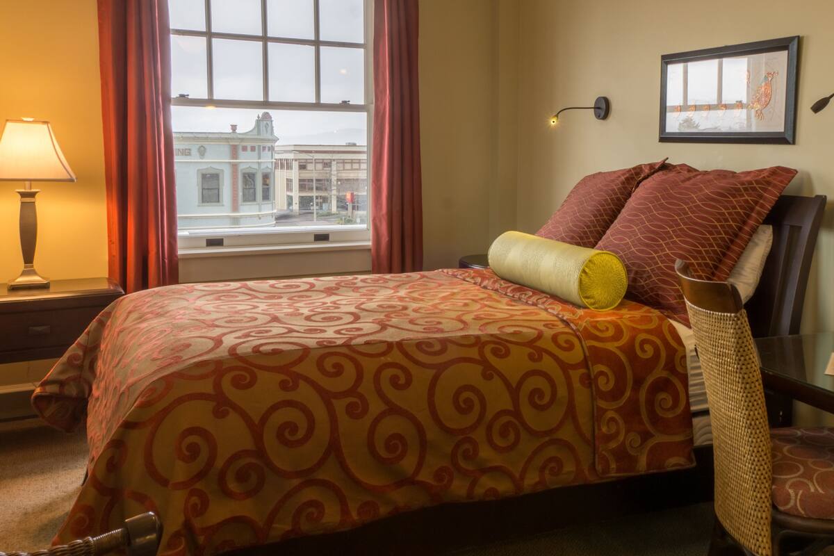 A hotel room with a large bed, a nightstand, a lamp, a chair, and a view of a building through the window.