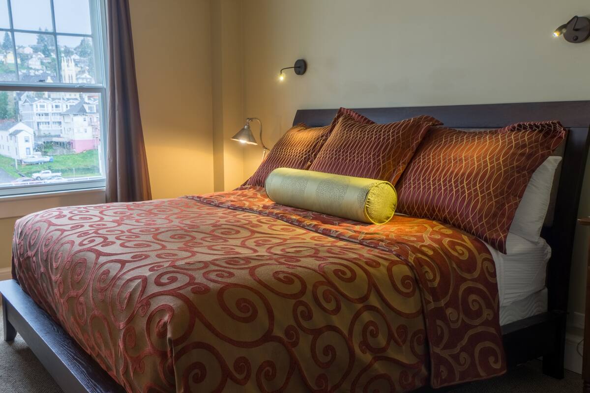 A neatly made bed with a patterned bedspread and pillows, a bedside lamp, and a view of buildings through a window.