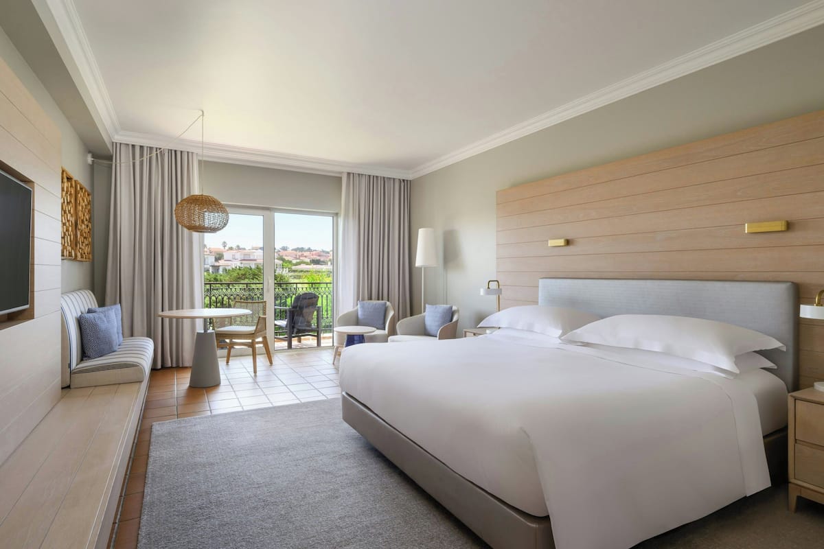 A hotel room with a large bed, a sofa, a small table, and a chair. There is a balcony with a view of the city.