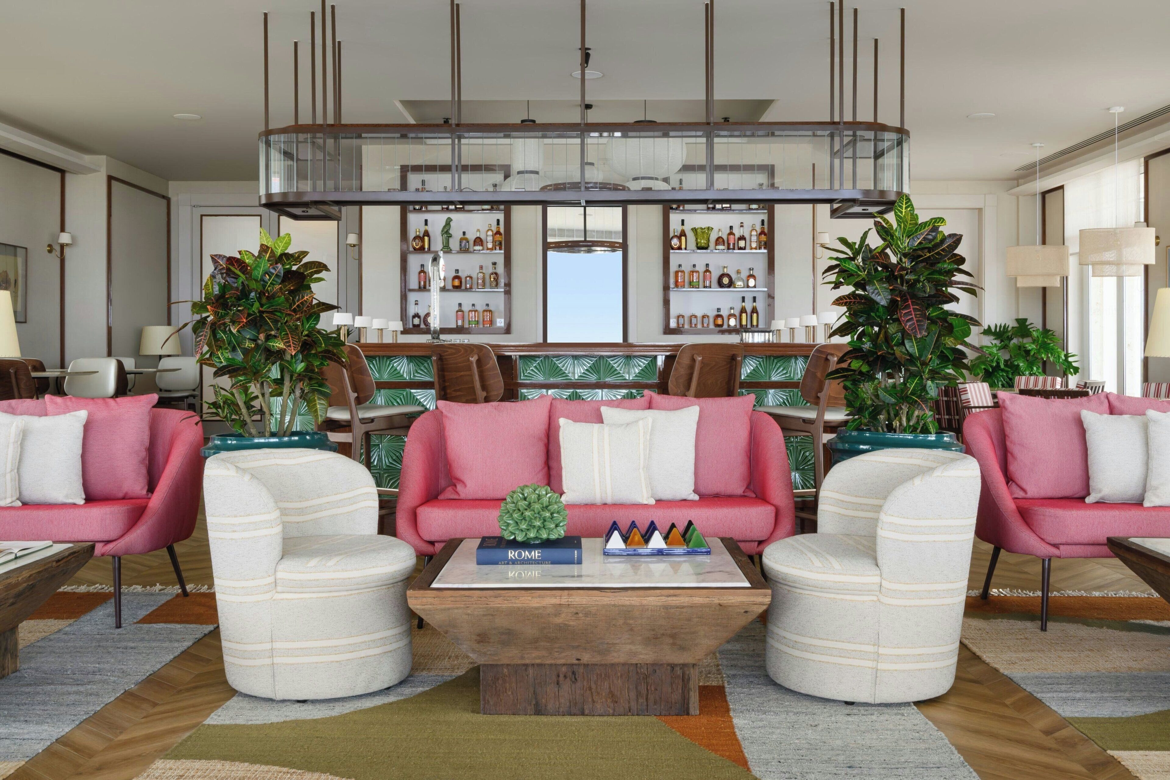 A modern living room with a central coffee table, two pink sofas, and a bar area with shelves of bottles.