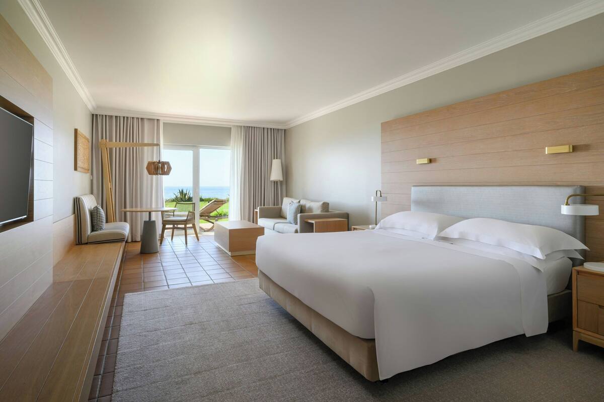 A modern hotel room with a large bed, a seating area with a table, and a view of the outdoors.