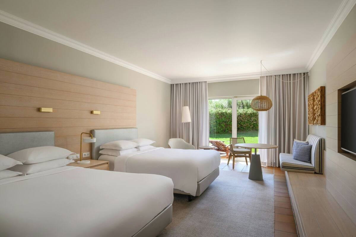 A hotel room with two beds, a sofa, a dining table, and a large window with curtains.