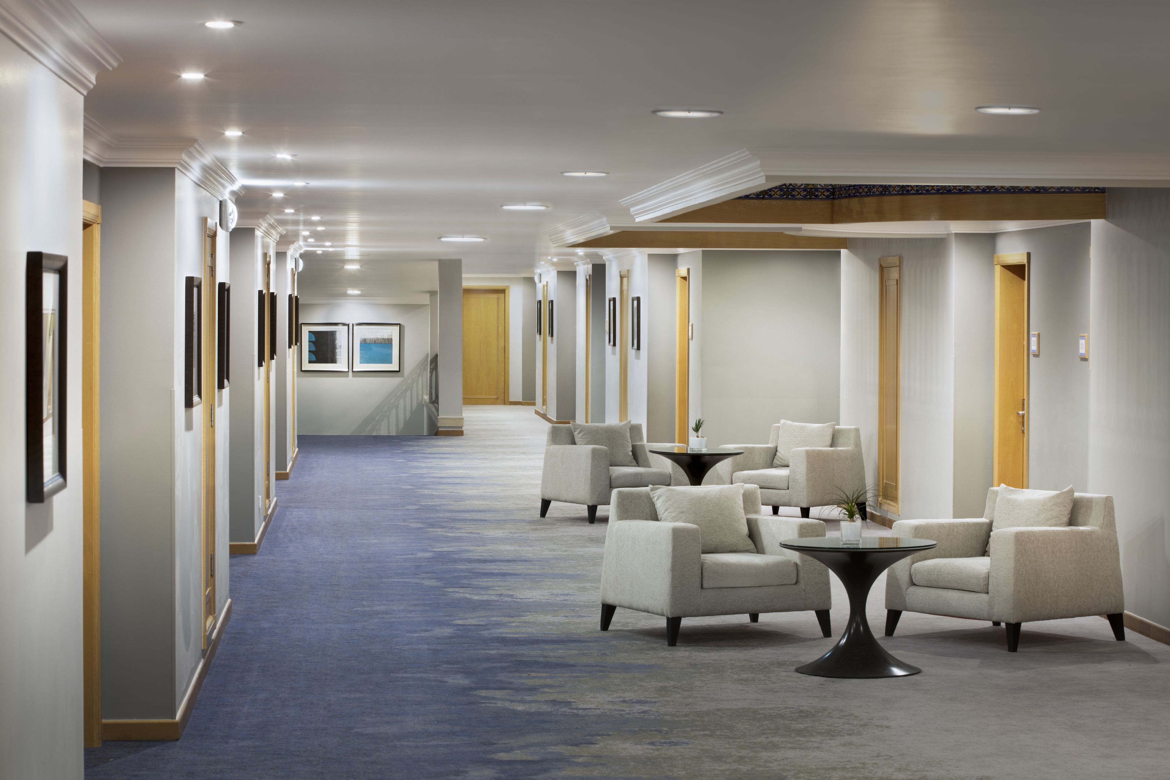 A hotel corridor with a blue carpet, multiple doors, framed pictures on the walls, and a seating area with a small table and chairs.
