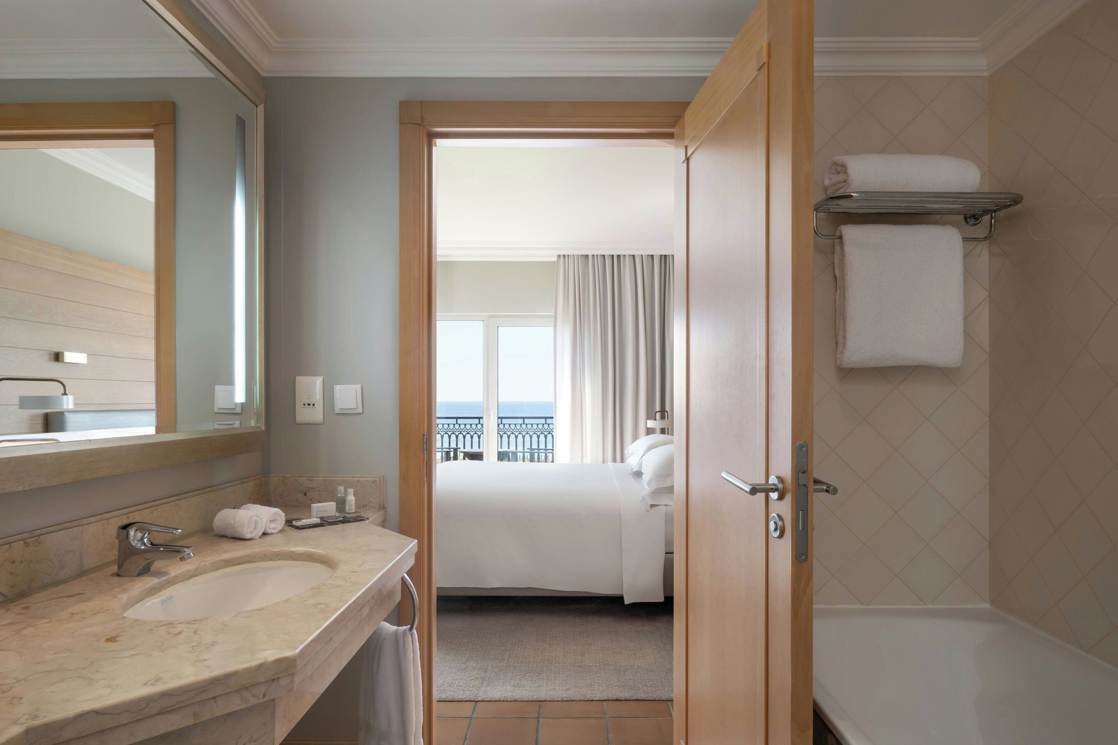 A hotel bathroom with a large sink, a mirror, a shower, and a view of the balcony.