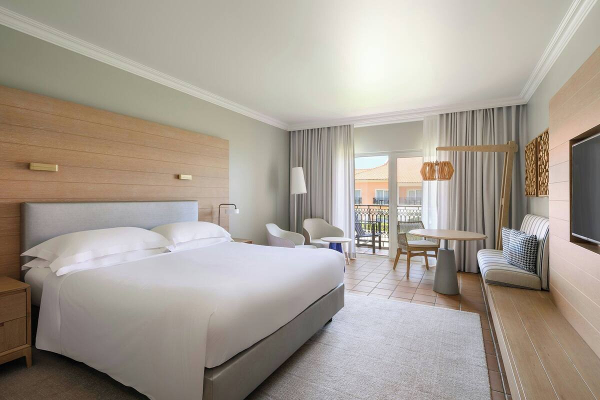 A hotel room with a large bed, a desk, a chair, a sofa, and a balcony with a view.