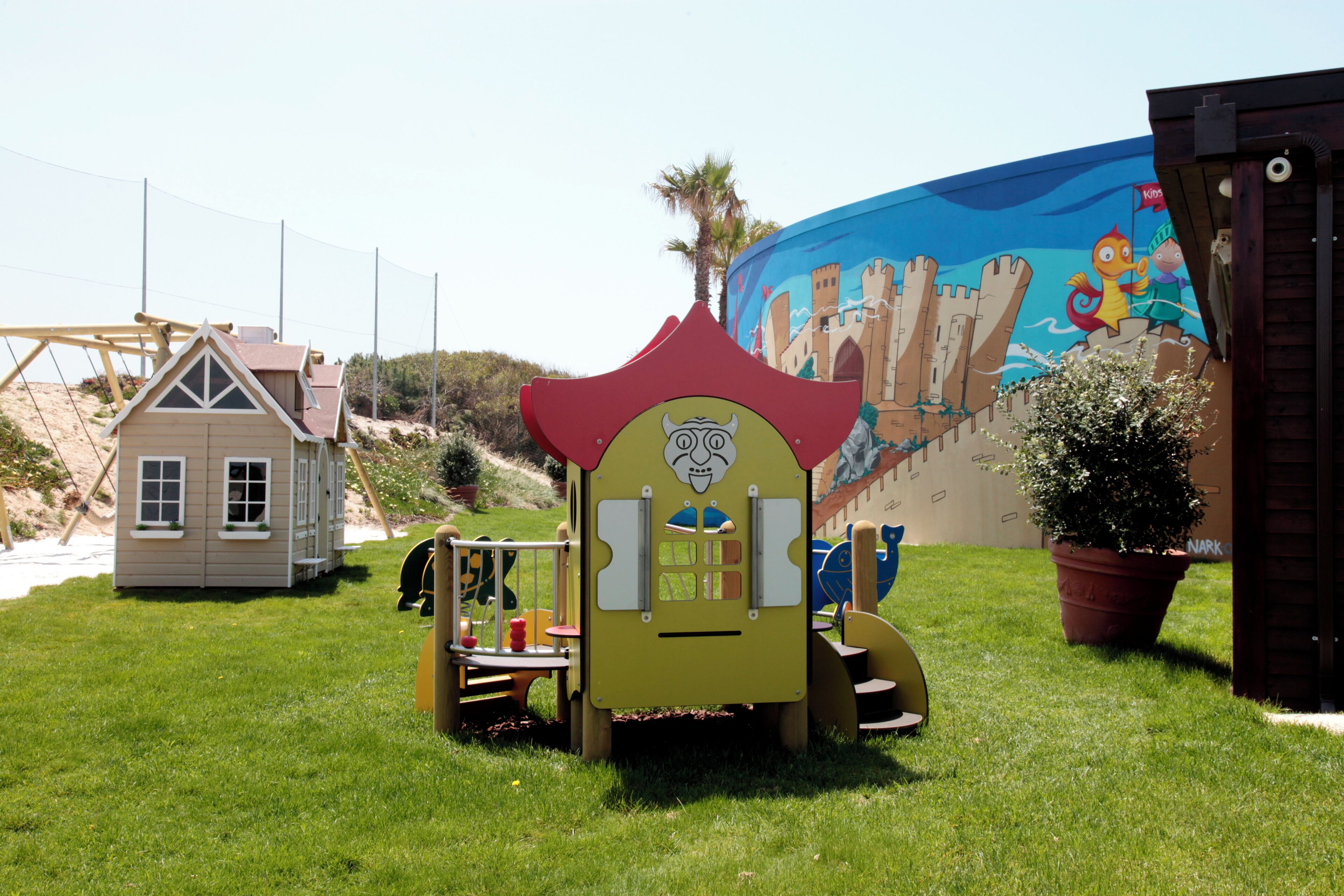 A playground with a slide and climbing structures, a small house-like playhouse, and a mural in the background.