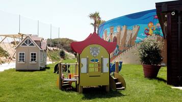 A playground with a slide and climbing structures, a small house-like playhouse, and a mural in the background.