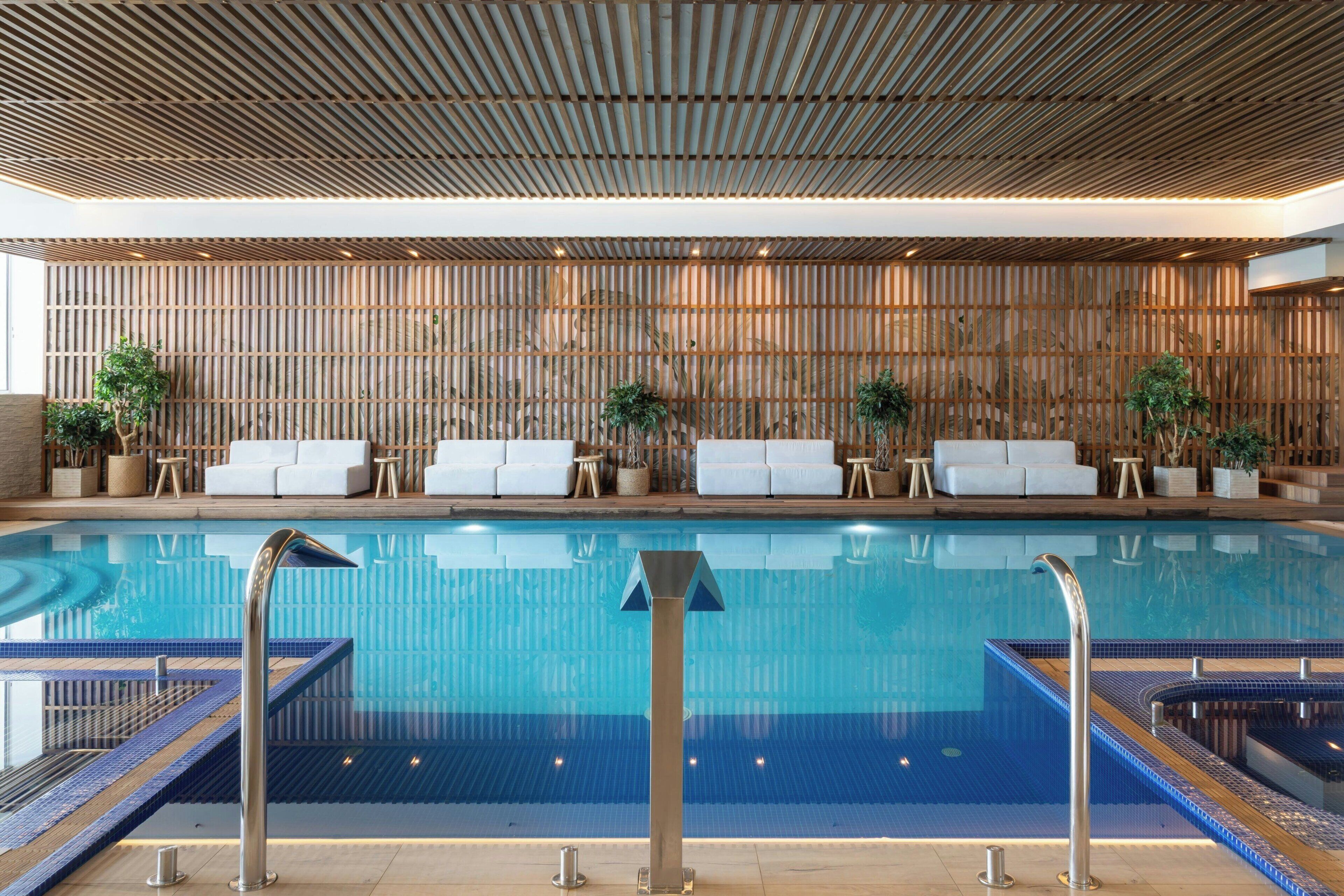 Una piscina interior moderna con agua azul cristalina, rodeada de sillones blancos y pequeñas mesas.