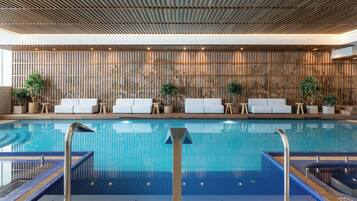 Una piscina interior moderna con agua azul cristalina, rodeada de sillones blancos y pequeñas mesas.