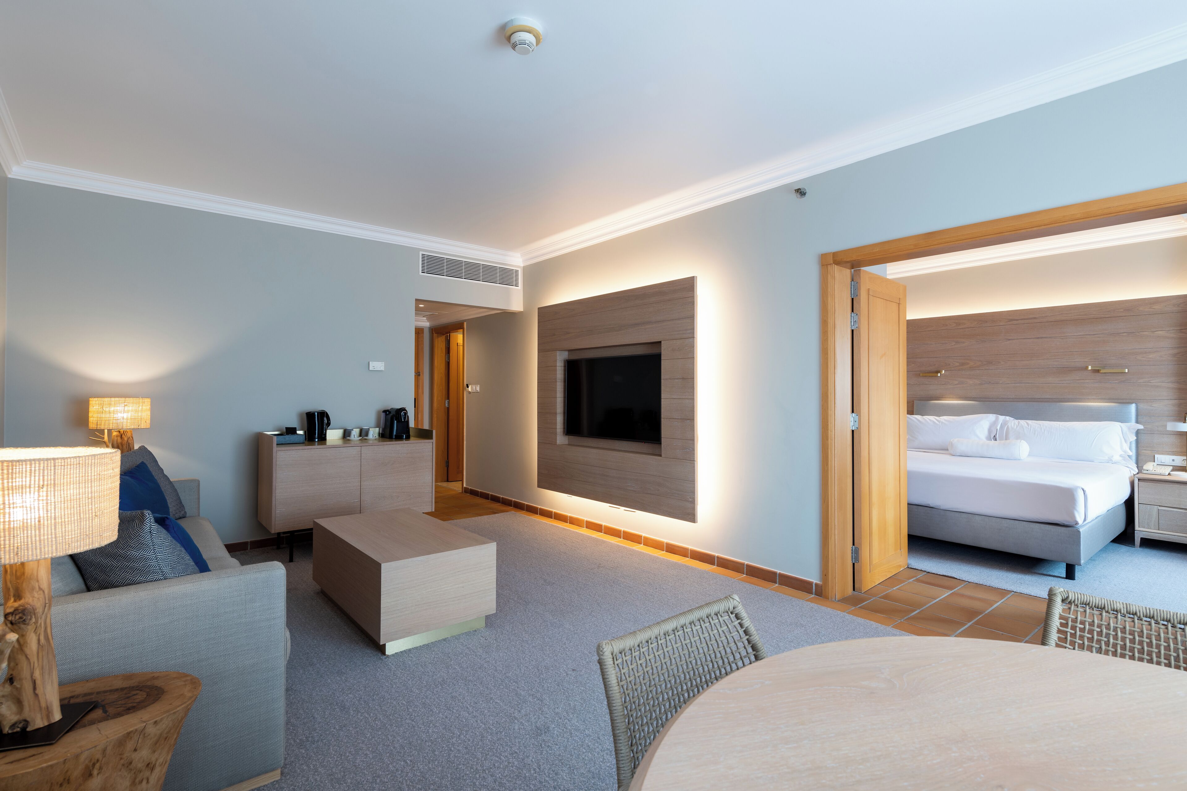 Une chambre d’hôtel moderne dotée d’un téléviseur à écran plat fixé au mur, d’un lit avec une tête de lit en bois, d’une table de chevet et d’un coin salon avec une table et des chaises.