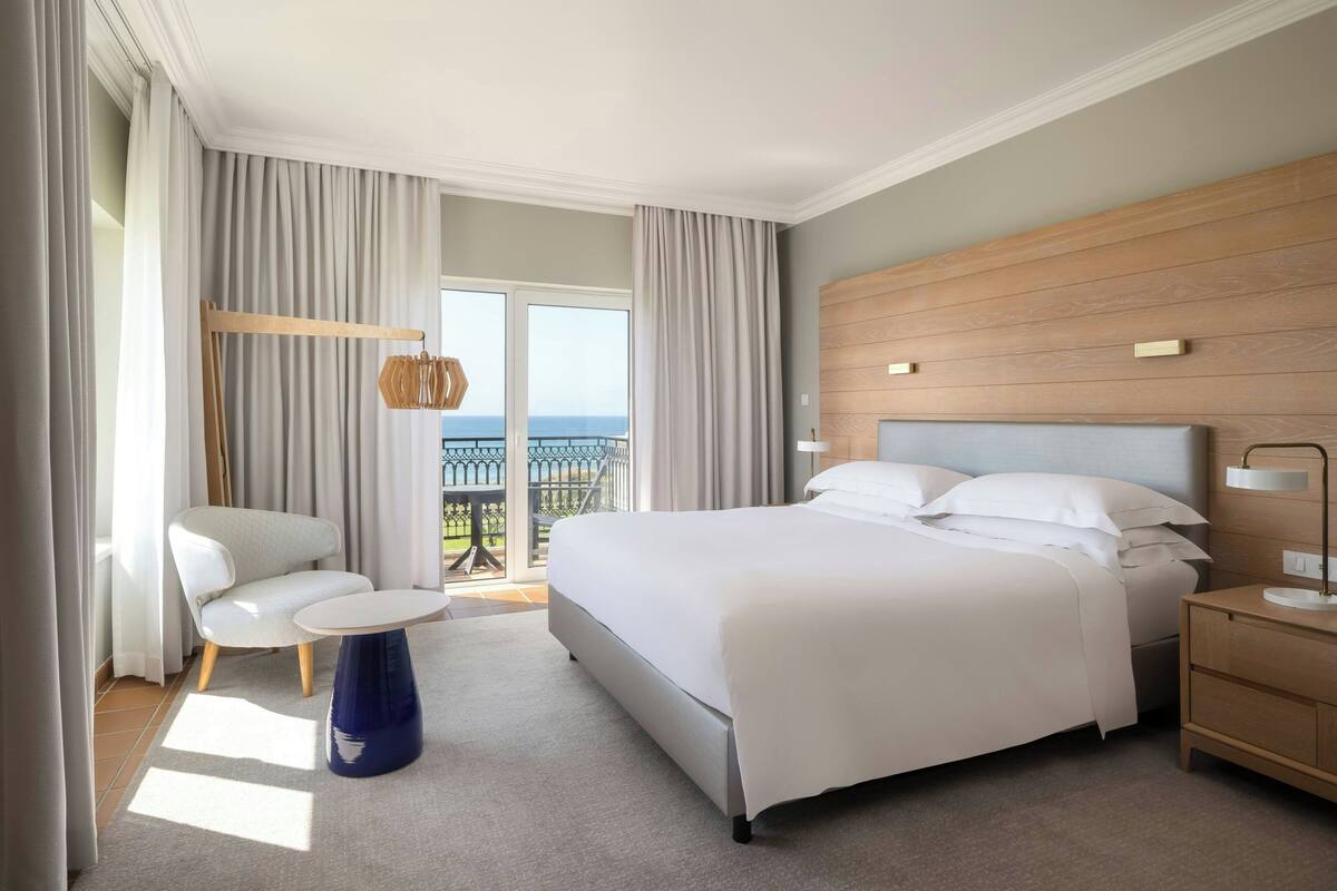 A hotel room with a large bed, a chair, a small table, and a nightstand. There is a balcony with a view of the sea.