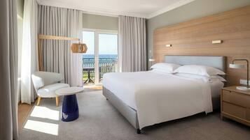 Quarto de hotel com uma cama grande, uma cadeira, uma mesinha e uma mesinha de cabeceira. Tem uma varanda com vista para o mar.