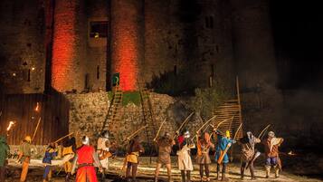 A medieval reenactment with participants in armor and costumes, set against a backdrop of a stone castle with red-lit towers.