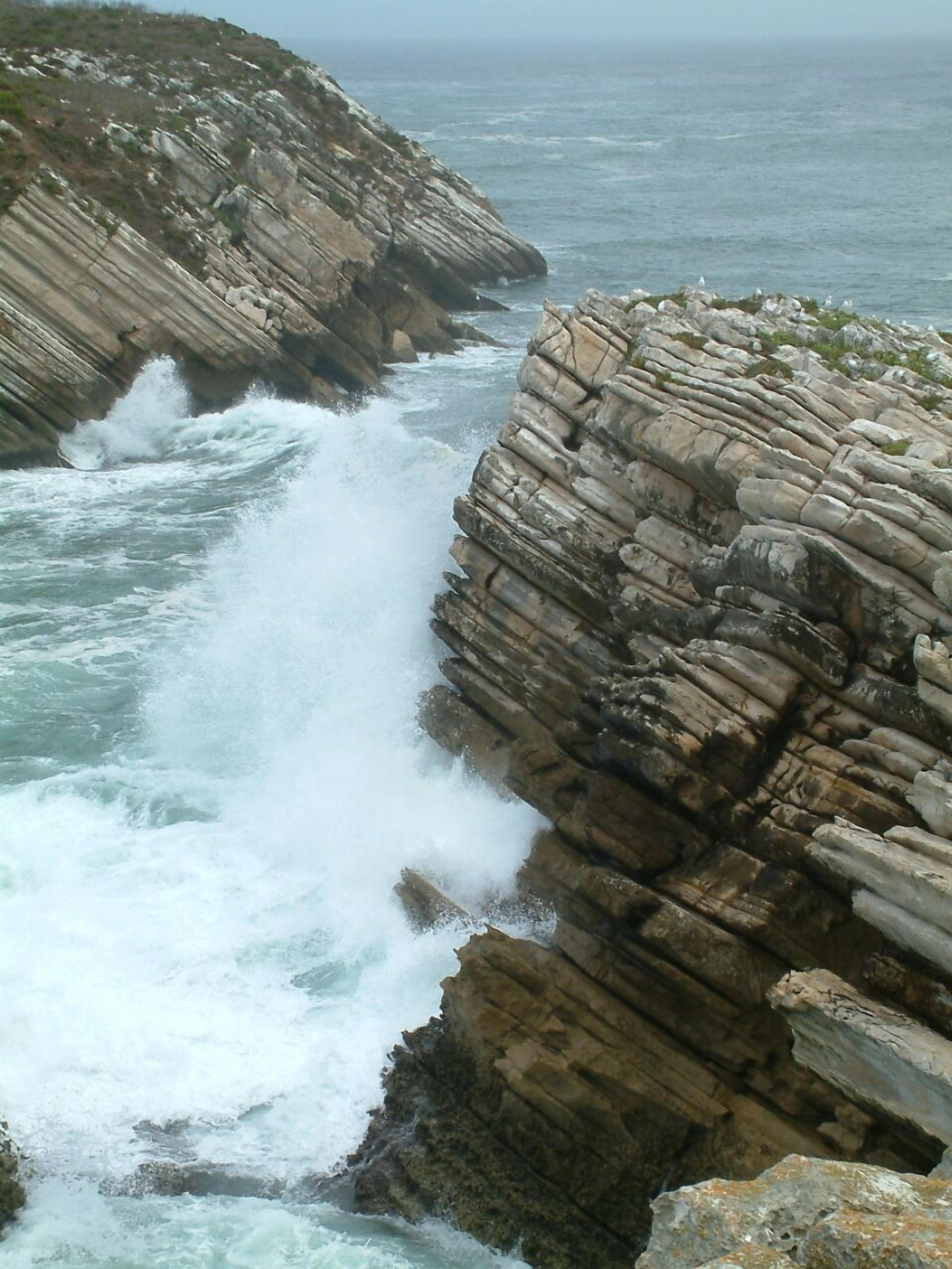 A coastal scene with a rocky cliff and waves crashing against it.