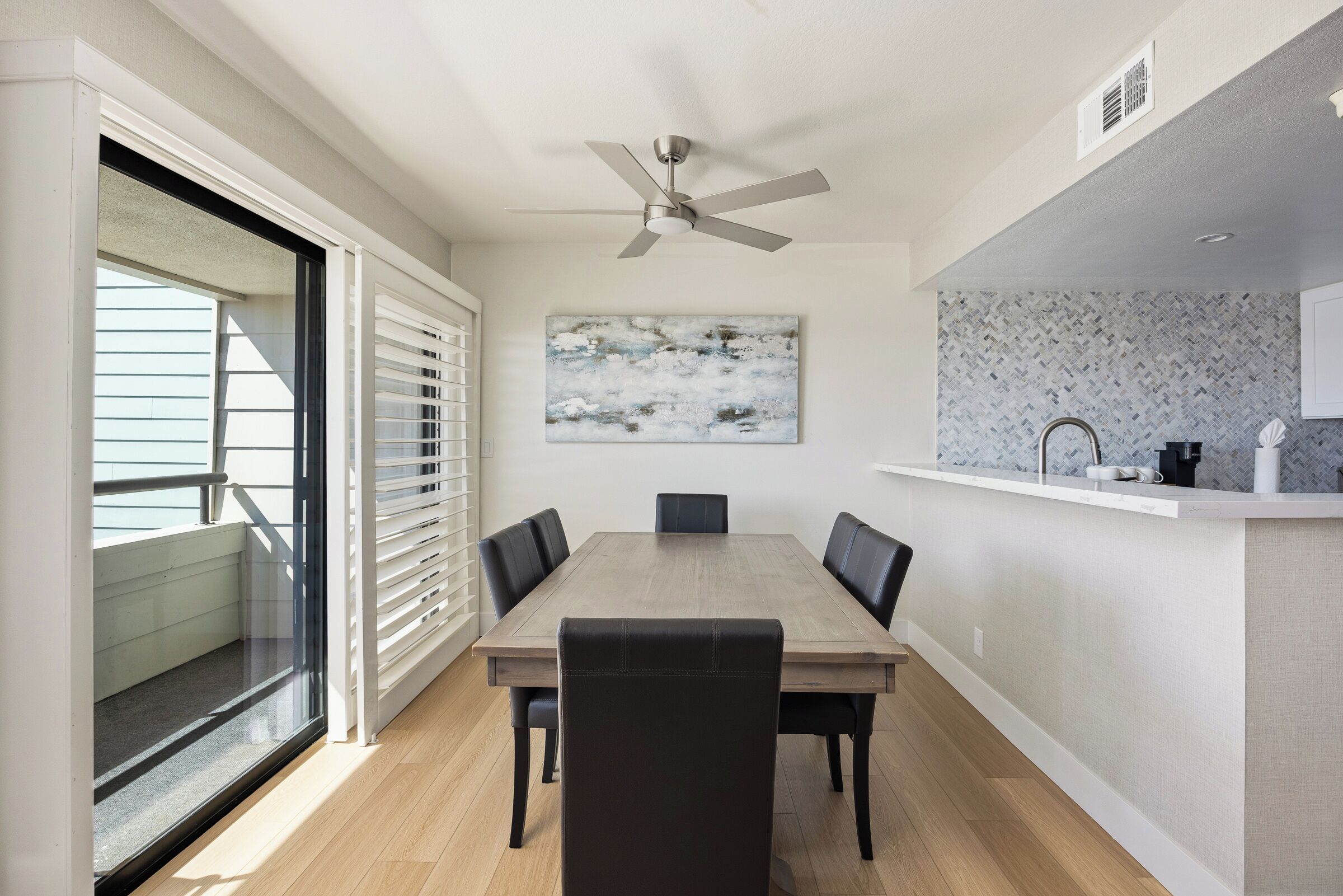 A dining area with a wooden table, black chairs, a ceiling fan, a large abstract painting, and a kitchen counter.