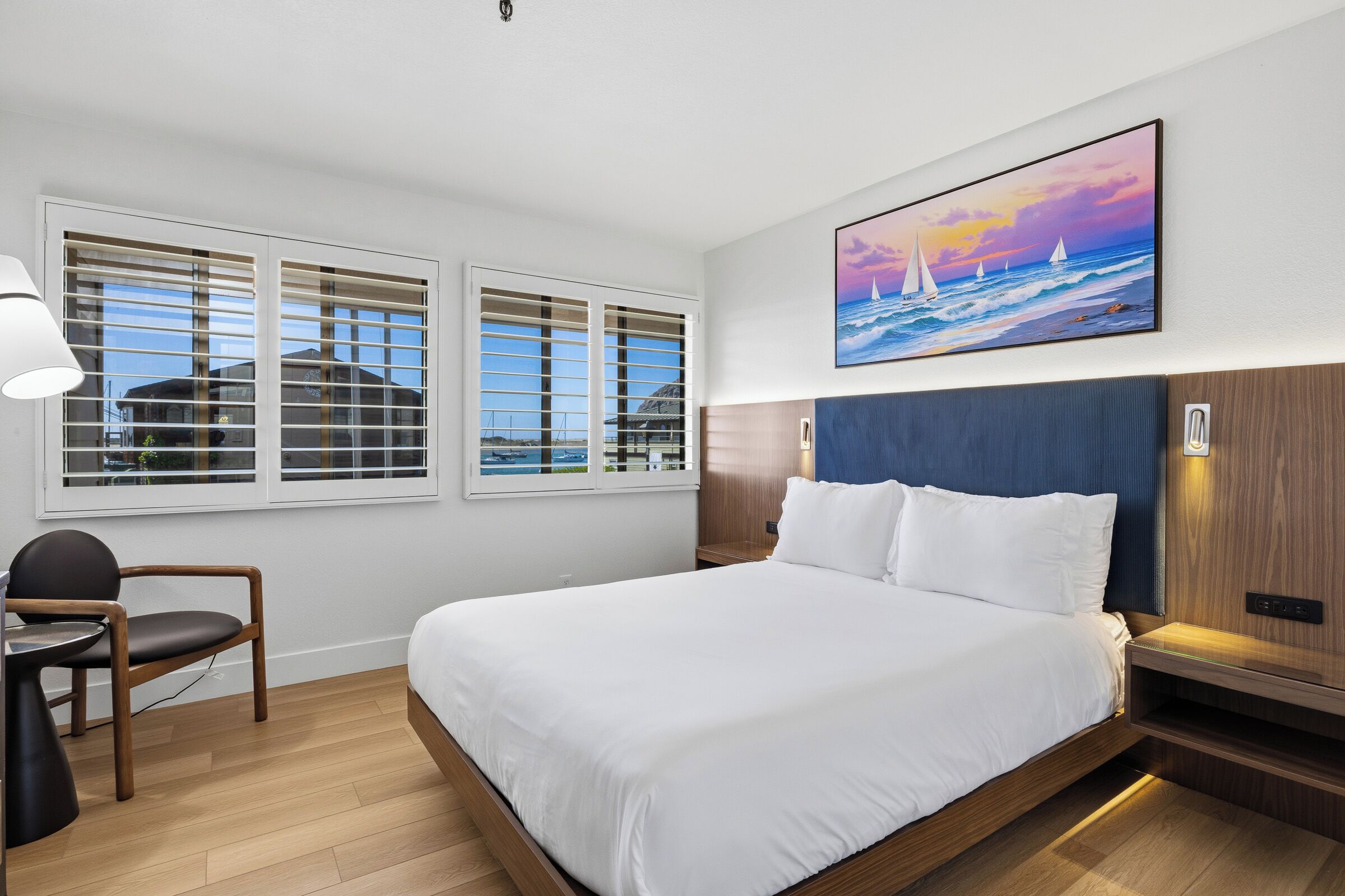A hotel room with a bed, a desk, a chair, and a painting of sailboats on the wall.