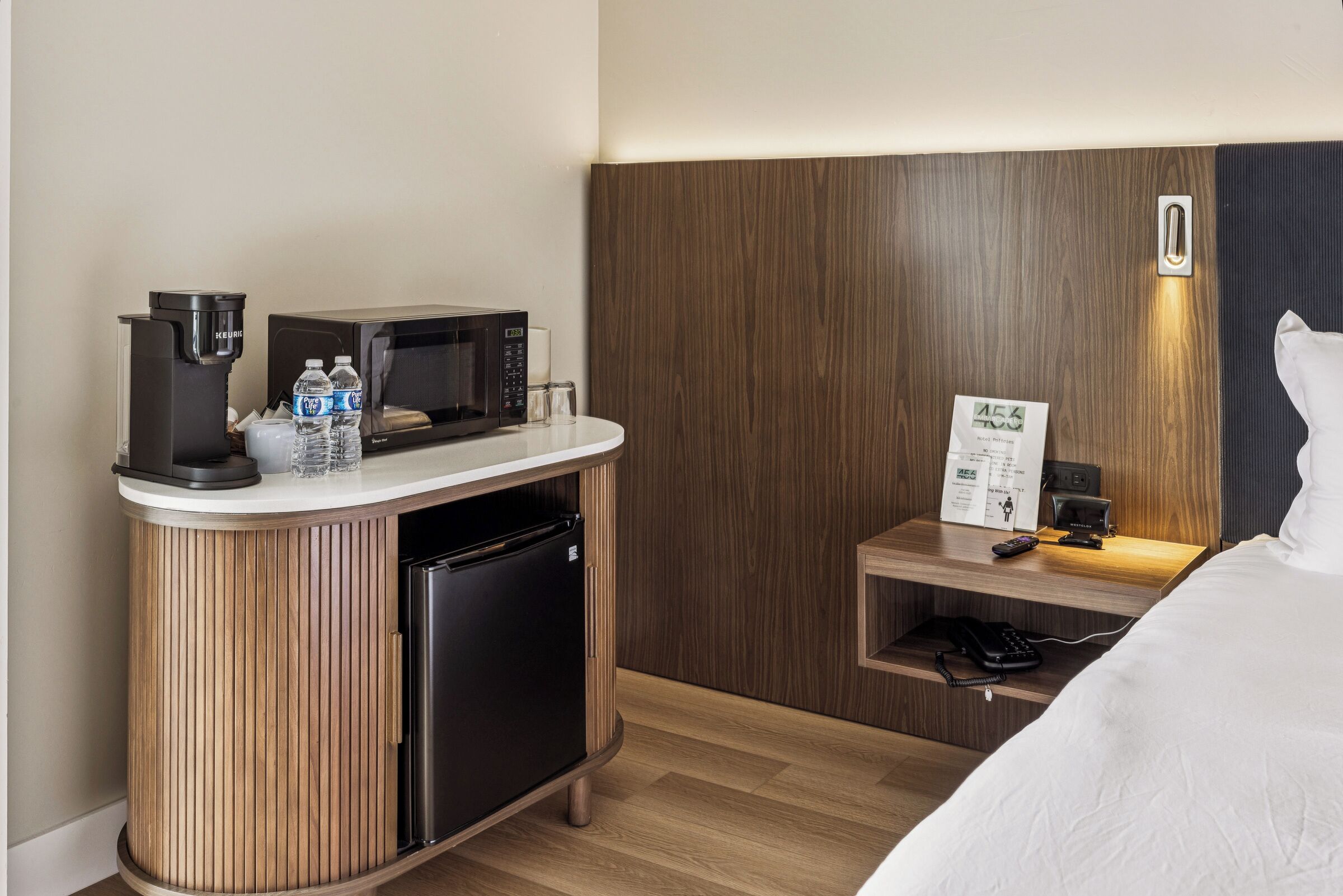 Habitación de hotel con un armario de madera, microondas, cafetera, refrigerador, mesita de noche con control remoto y una cama.