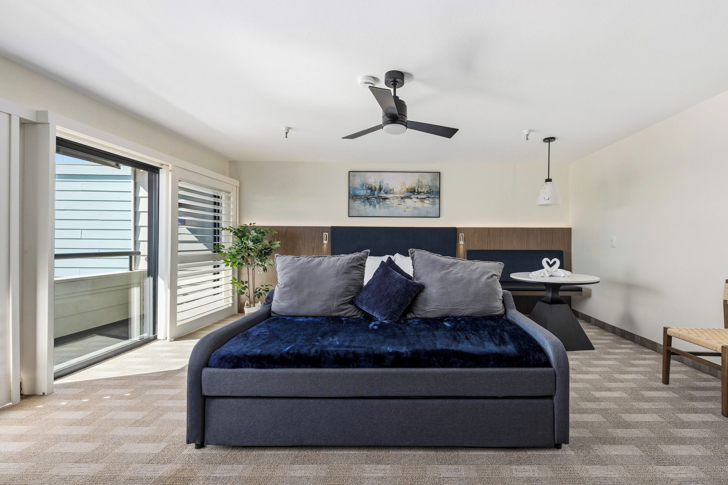 A modern bedroom with a bed, a ceiling fan, a painting, a small table, and a chair.
