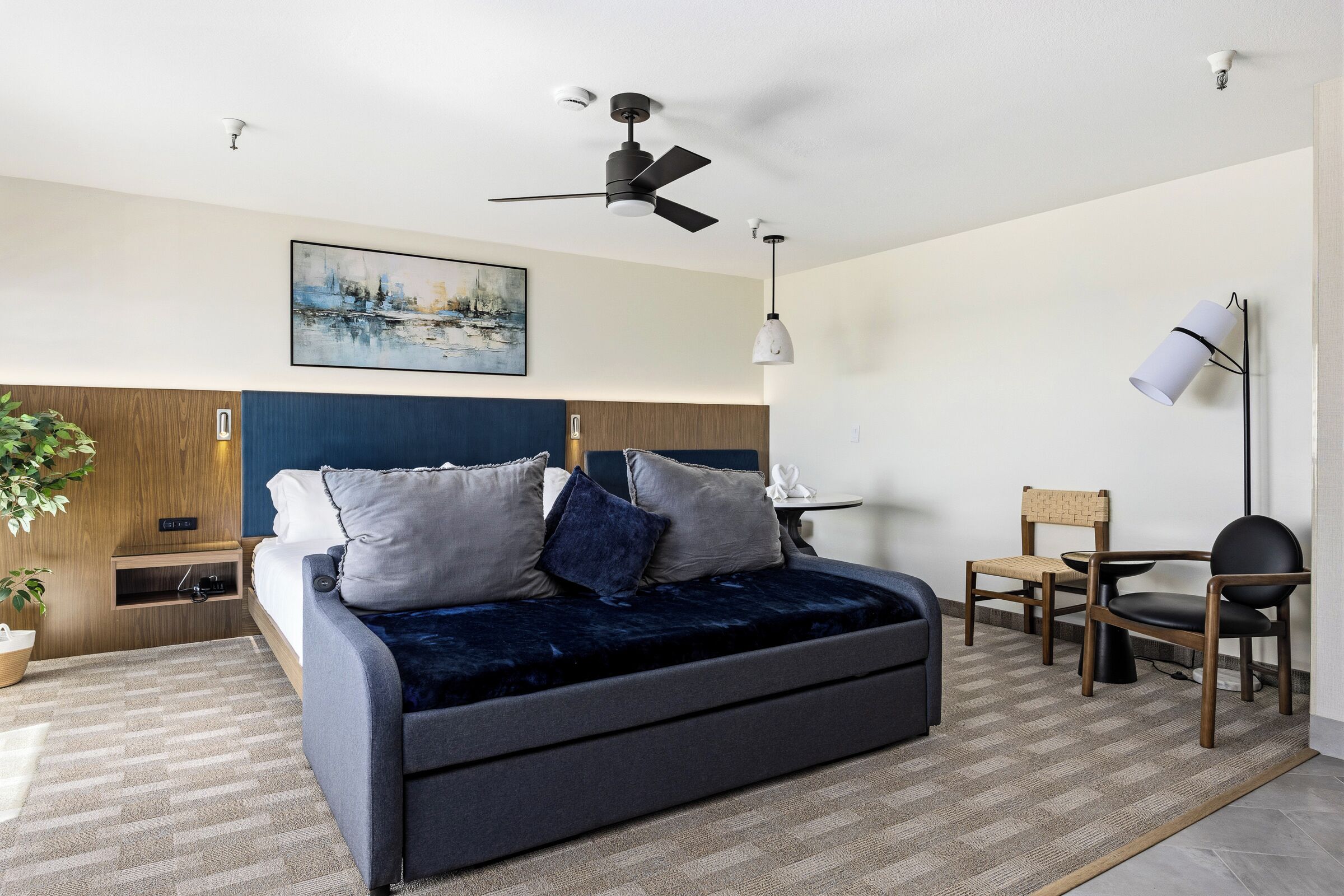 Una habitación de hotel moderna con cama, escritorio, silla, ventilador de techo y un cuadro en la pared.