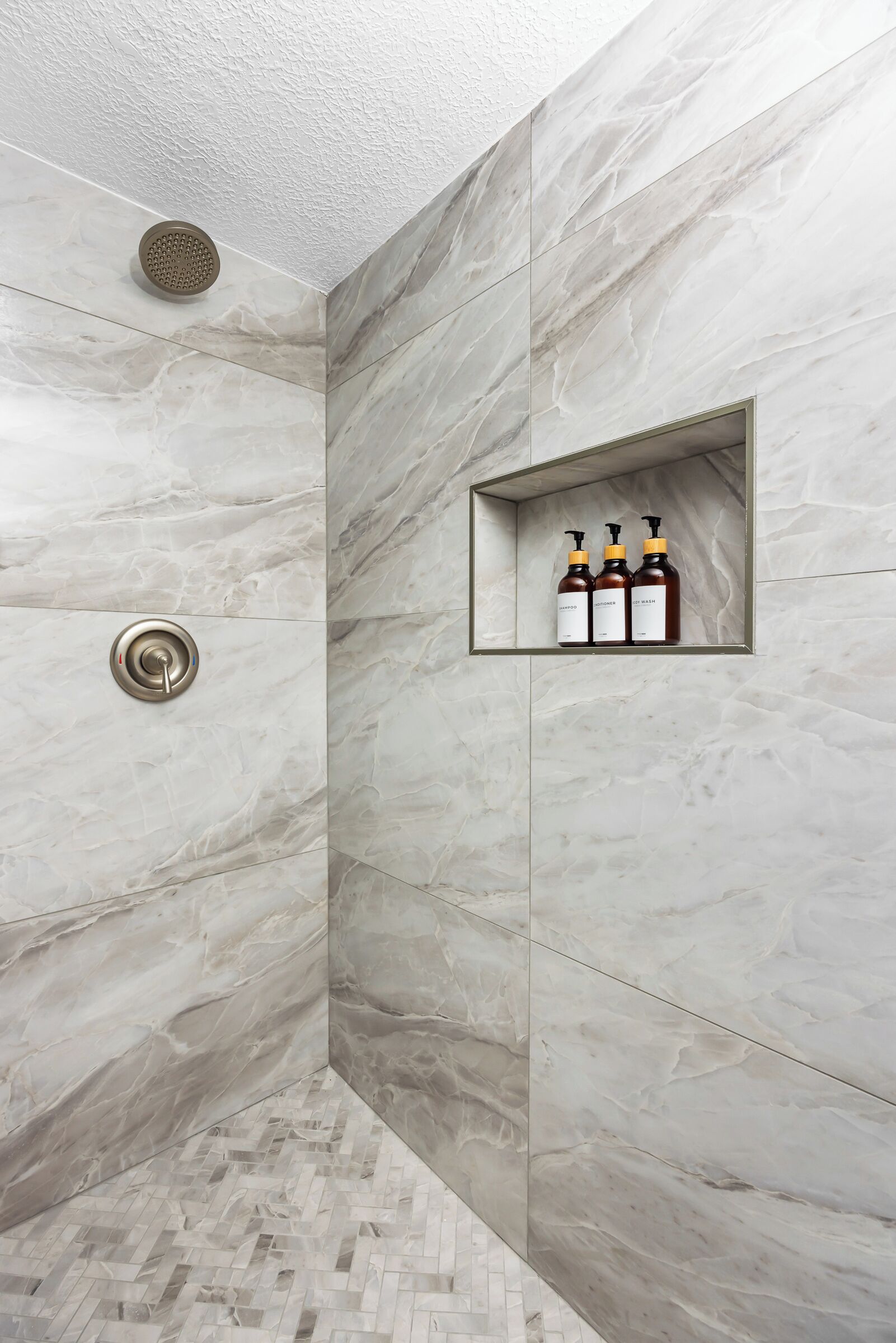 Une douche avec un mur en marbre, une étagère intégrée où sont rangées des bouteilles, et un sol orné d’un motif en chevrons.