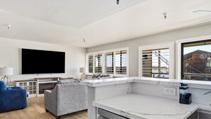 A modern living room with a flat-screen TV, grey sofa, and a kitchen area with a marble countertop and window with blinds. - 456 Embarcadero Inn & Suites (Morro Bay)