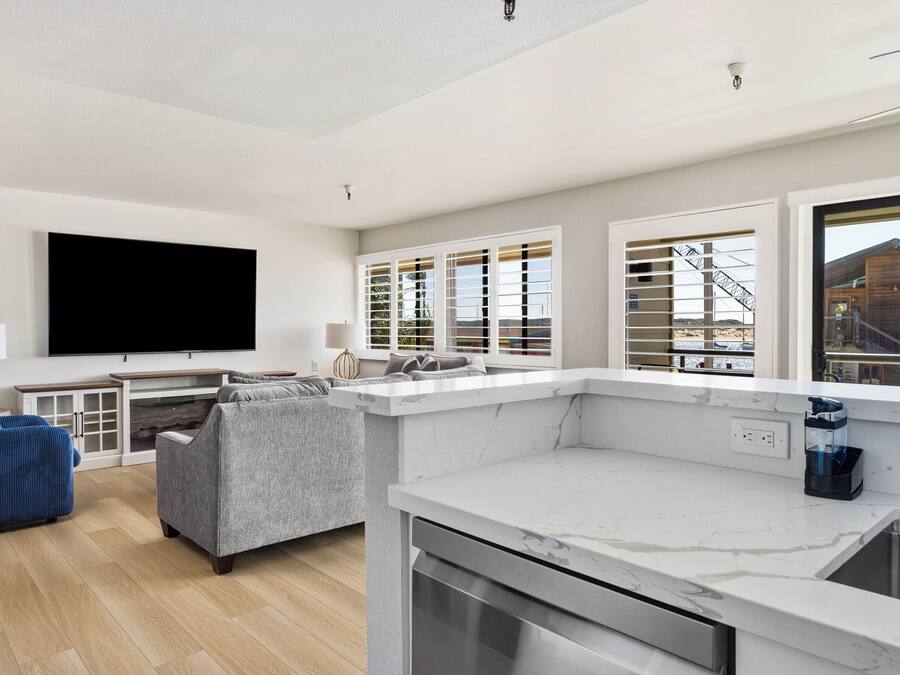 Un salón moderno con televisor de pantalla plana, sofá gris y una cocina con encimera de mármol y ventana con persianas.