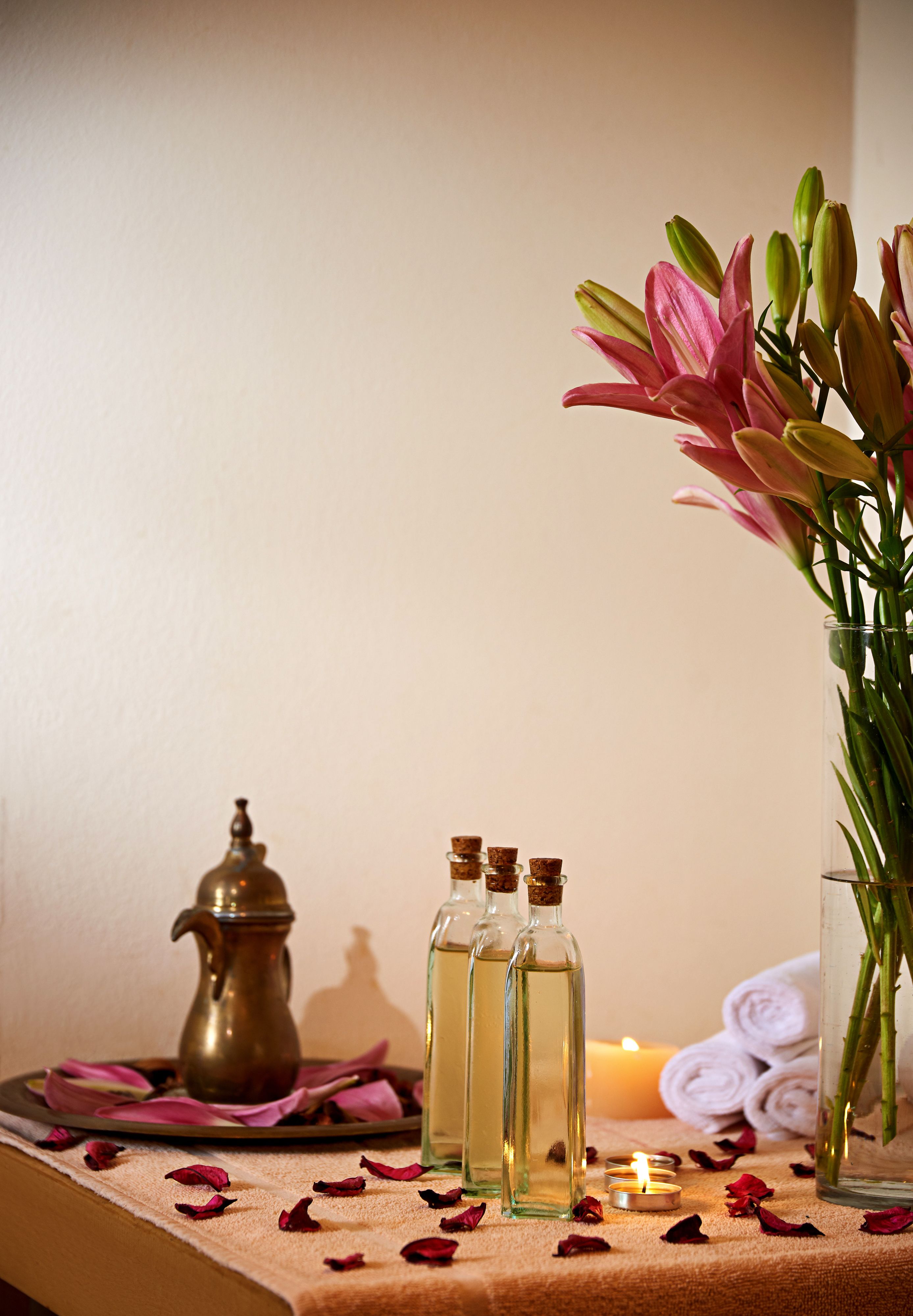 Uma mesa com um jarro de latão, três garrafas de vidro, uma vela acesa, um vaso com flores e toalhas enroladas.
