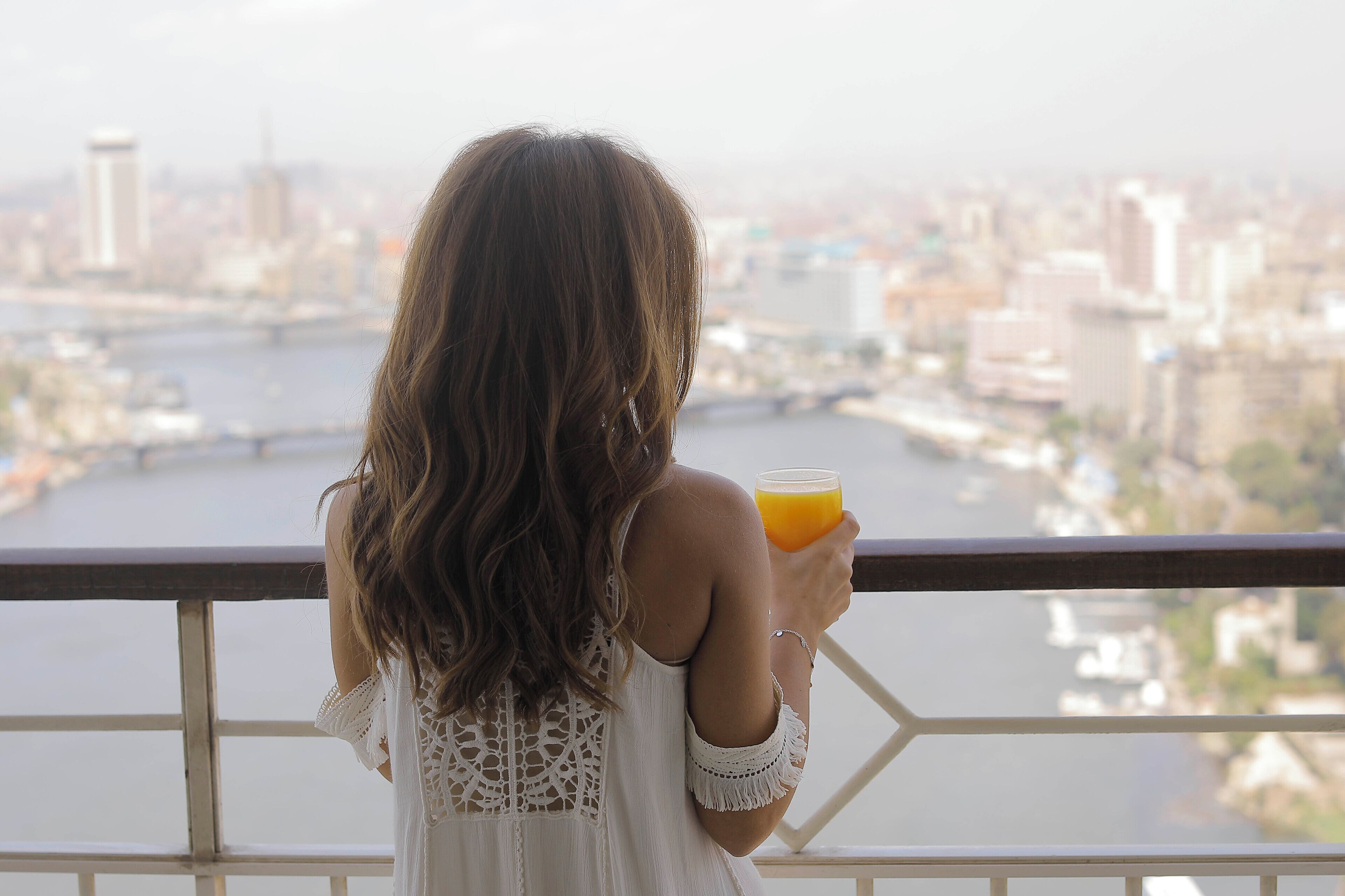 Una persona sostiene un vaso de jugo de naranja, con vistas a un paisaje urbano que incluye un río y edificios.