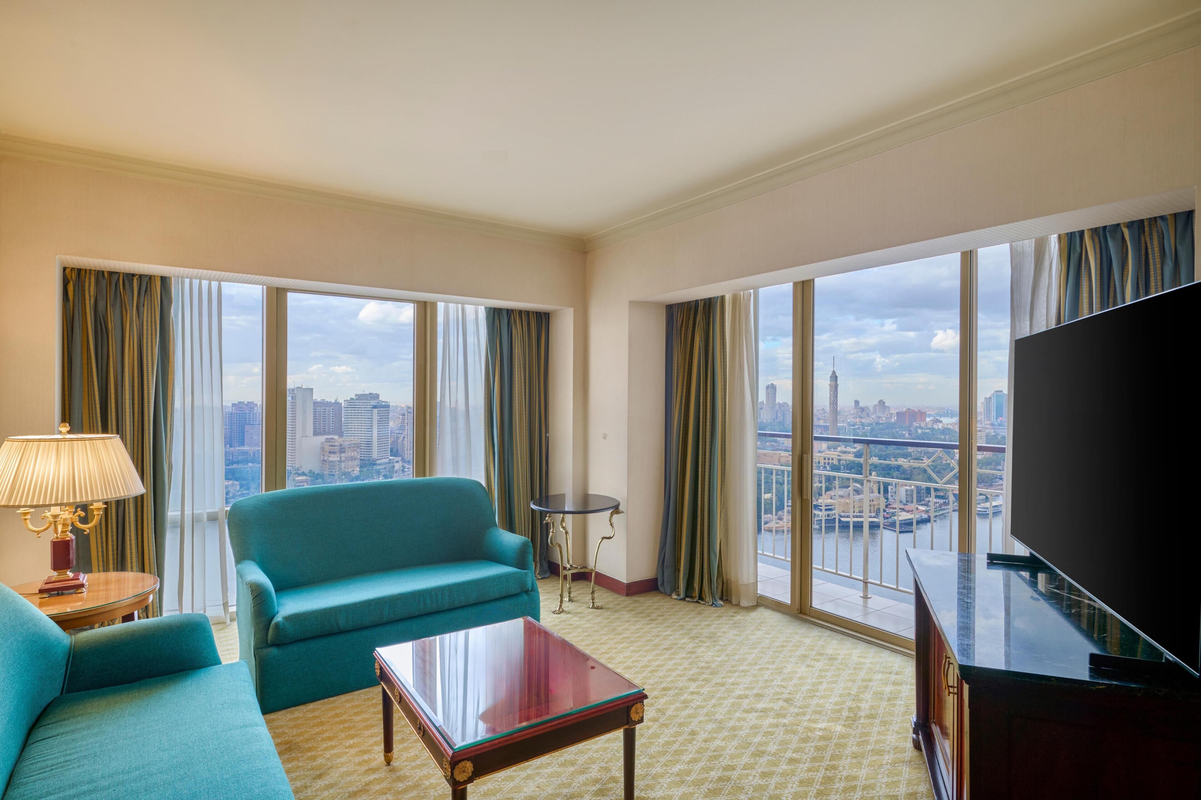 Une chambre d’hôtel avec une grande fenêtre, un canapé de couleur turquoise, une petite table ronde et un téléviseur.