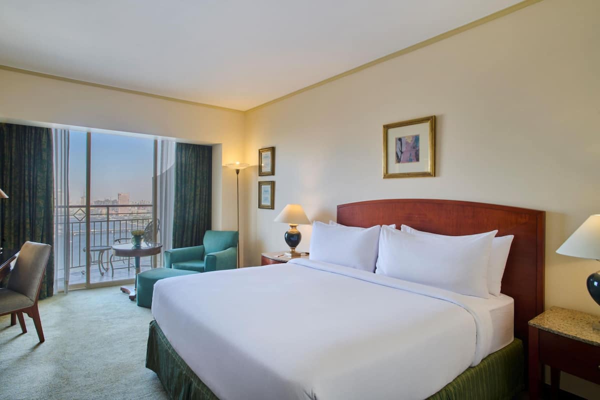 A hotel room with a large bed, a chair, a small table, and a view through a large window.