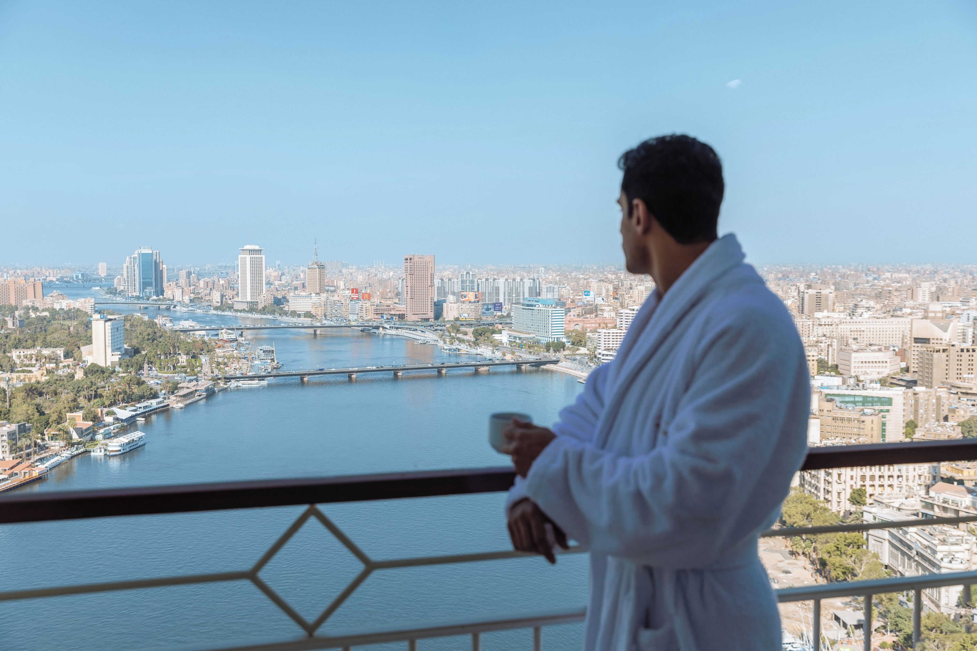 Un hombre con bata blanca de pie junto a un balcón con vistas a un paisaje urbano que incluye un río y puentes.