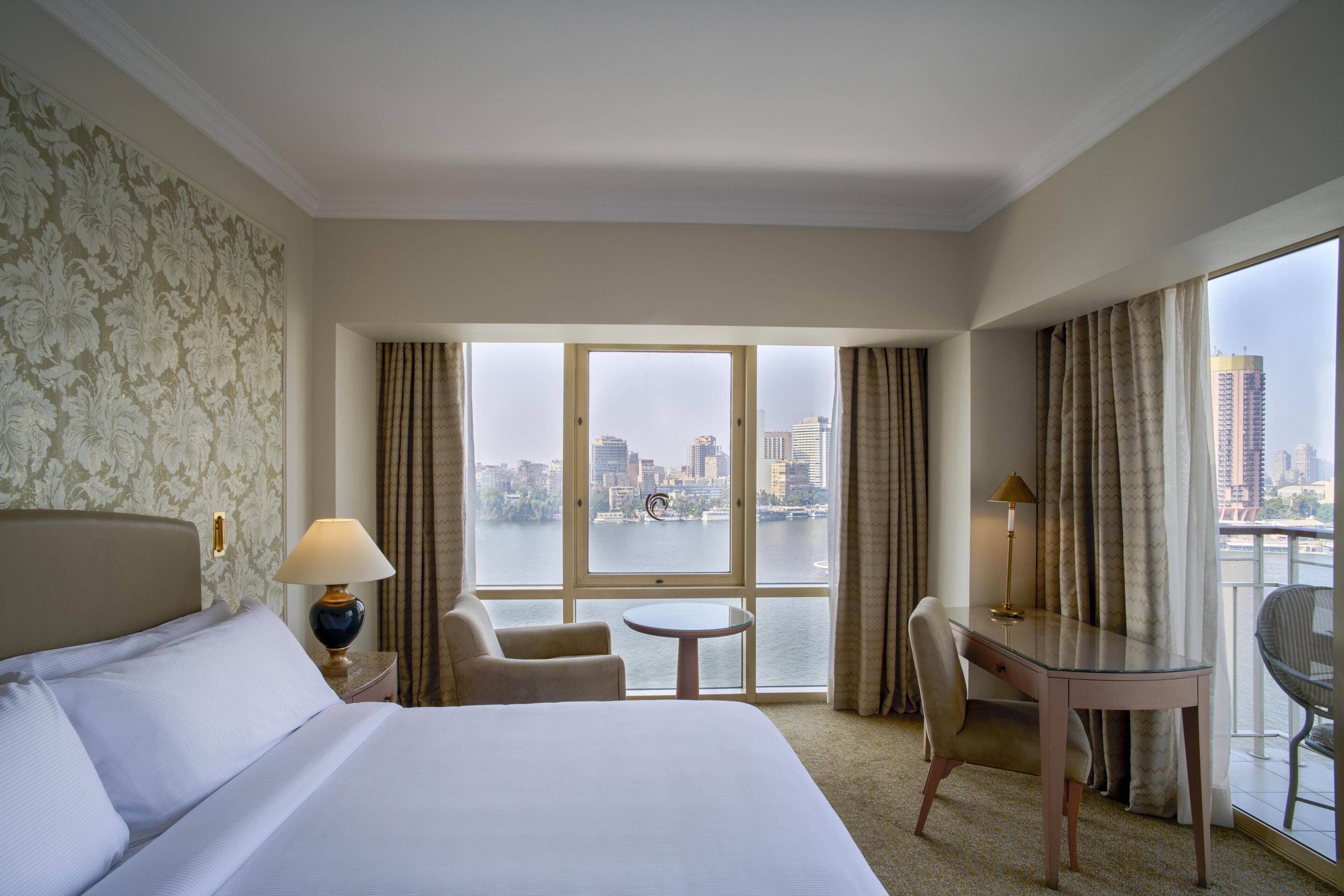 A hotel room with a large bed, a desk, a chair, a small table, and a view of a cityscape through large windows.