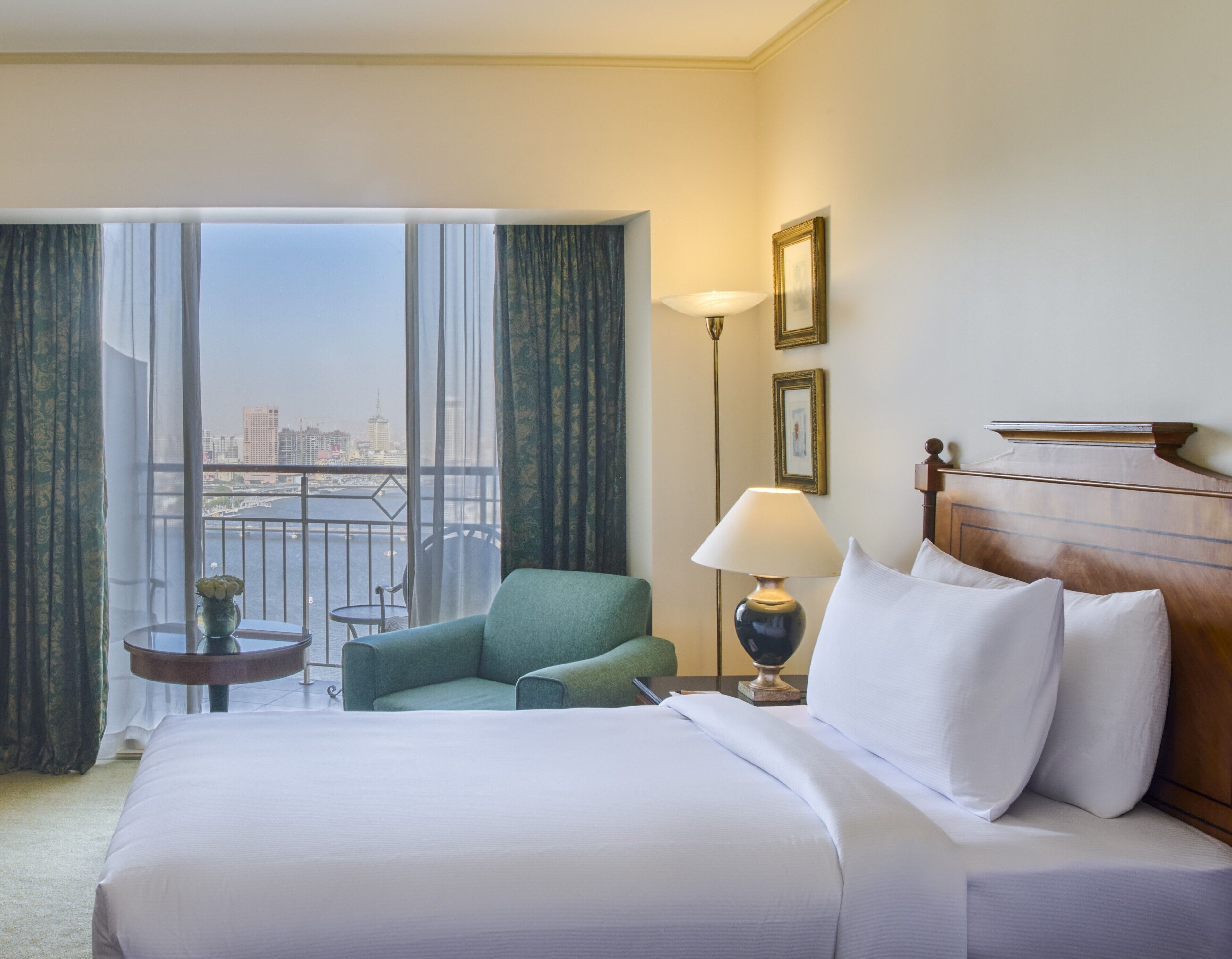 A hotel room with a large bed, a green armchair, a small table with a vase of flowers, a lamp, and a view of a cityscape through large windows.