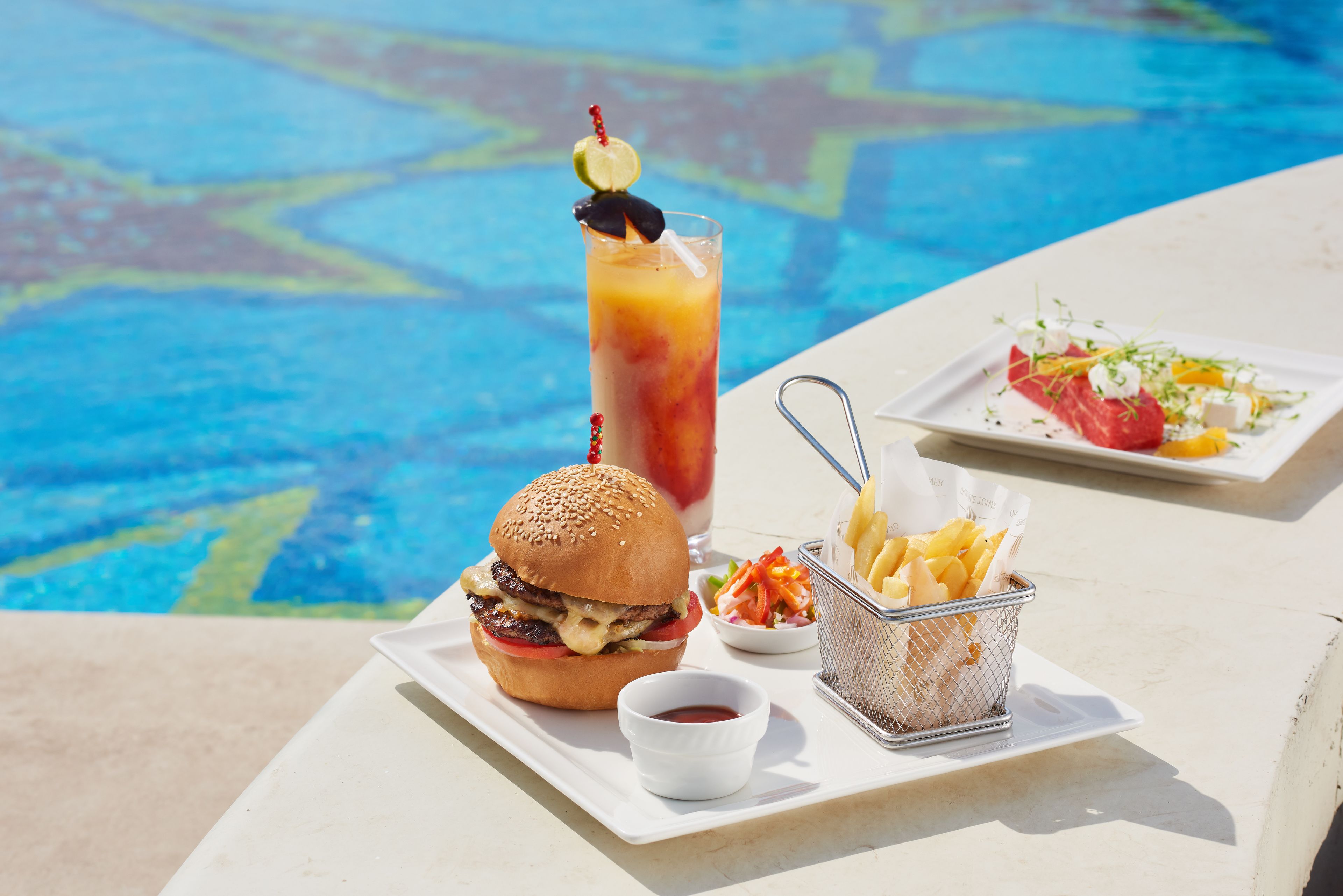 A meal with a burger, fries, a salad, and a drink by a poolside.