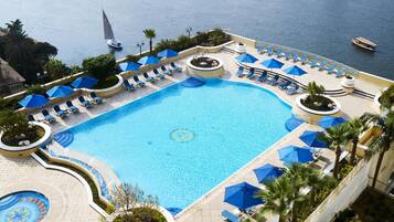 A large swimming pool with sun loungers and umbrellas, surrounded by palm trees and a view of the sea.