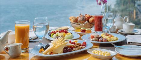 A breakfast table with various food items, a pitcher, and a vase with flowers, overlooking a body of water with buildings in the background.